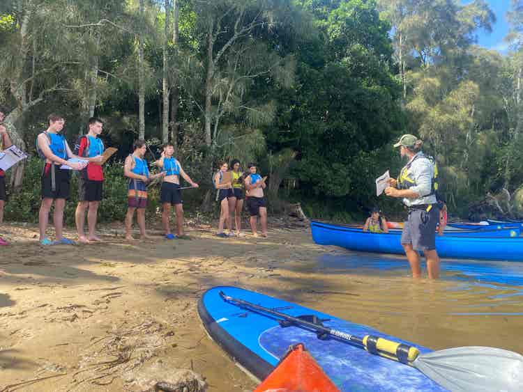 Teacher standing in shallow water next to canoe, addressing a group of students on the shore